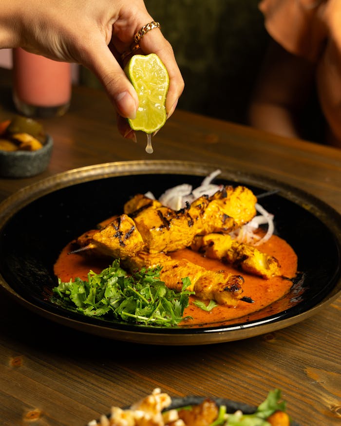 A hand squeezes lemon over tandoori chicken on a sauce and garnished with cilantro.